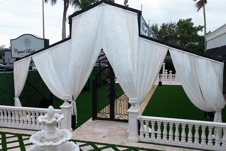 Elegant white fabric draping at the entrance of a wedding tent featuring a wrought iron gate and stone fountain.
