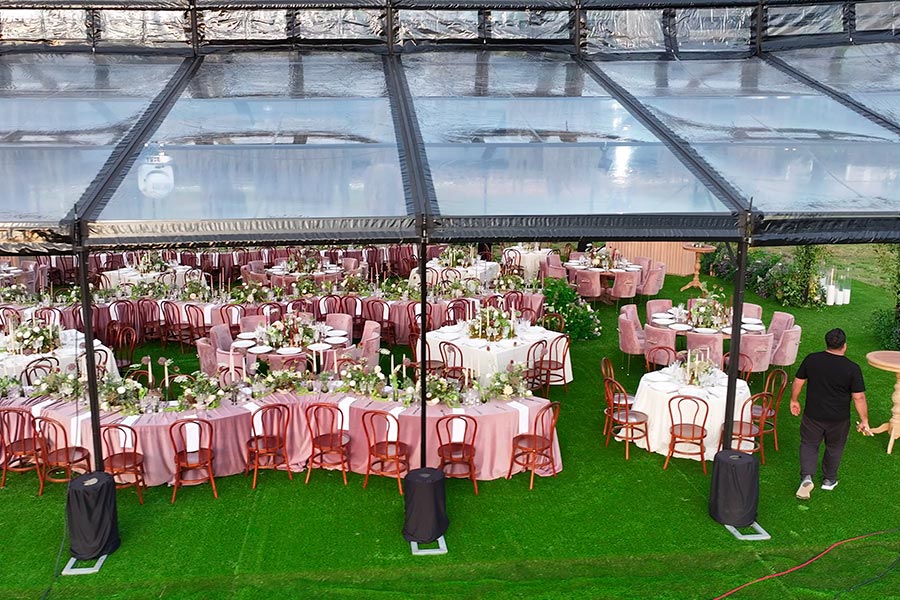 Large clear-top party tent with a black metal frame and transparent roof panels on a green lawn.