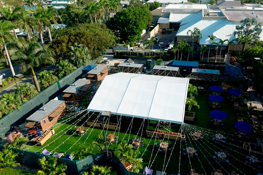 Aerial view of a large white event tent with string lights and outdoor dining tables on a green lawn.