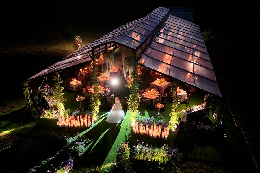 Night view of an illuminated clear-roof wedding tent with warm candlelight and hanging floral decor.