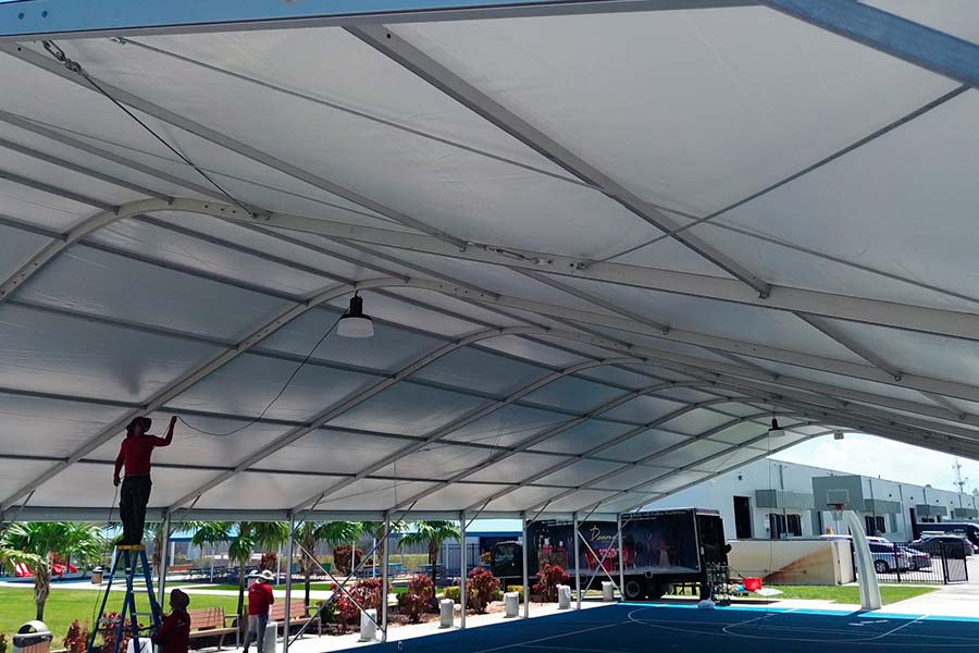 Worker installing lighting under a large white metal frame event tent on a basketball court.
