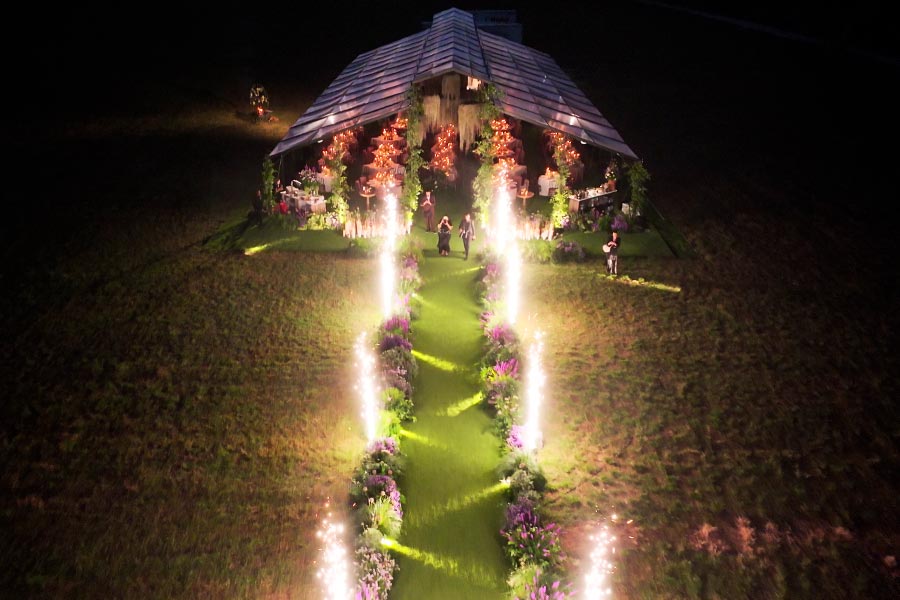 Aerial view of a lit clear-top wedding tent at night with a grand floral walkway and pyrotechnics.