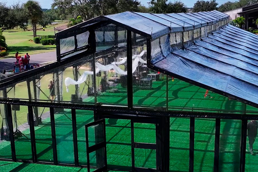 Large clear top frame wedding tent with black structure, glass doors, and green turf flooring on a sunny lawn.