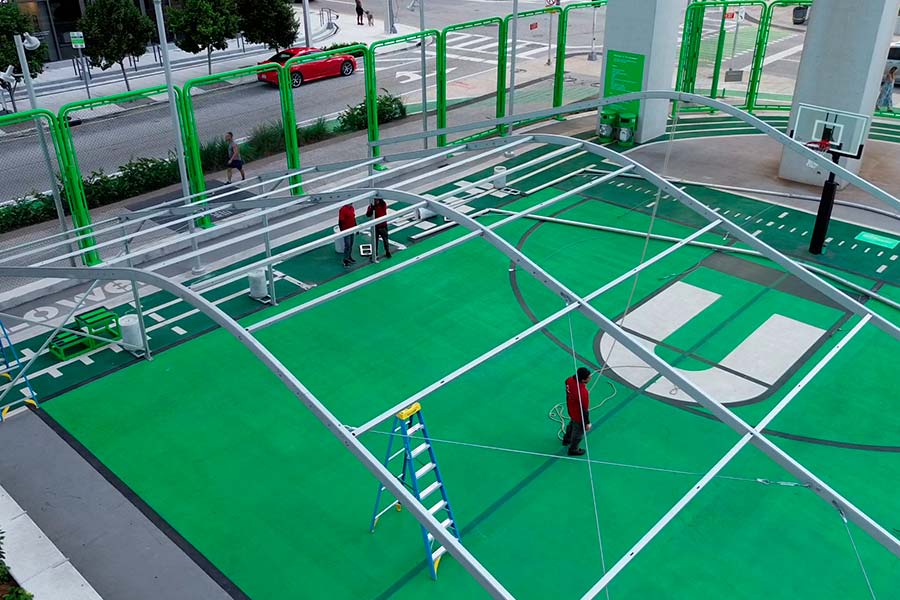 Aerial view of technicians securing the metal skeletal frame of a large event tent on a green court.