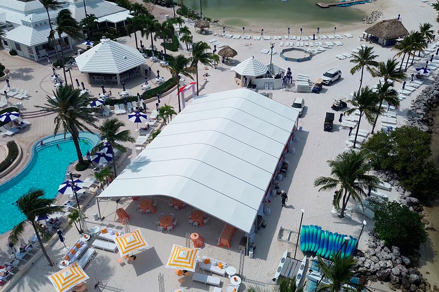 Aerial view of a large white frame tent on a beach resort with pool and tropical palm trees.