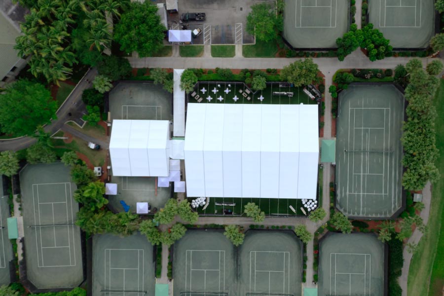 Top-down aerial view of a professional white event tent installation on a green tennis court surface.