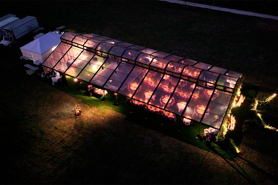 Aerial night view of a glowing clear-top wedding tent with warm ambient lighting and elegant dinner tables.