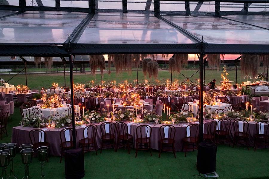 Elegant evening wedding reception under a clear-top tent with purple linens, candles, and floral arrangements.