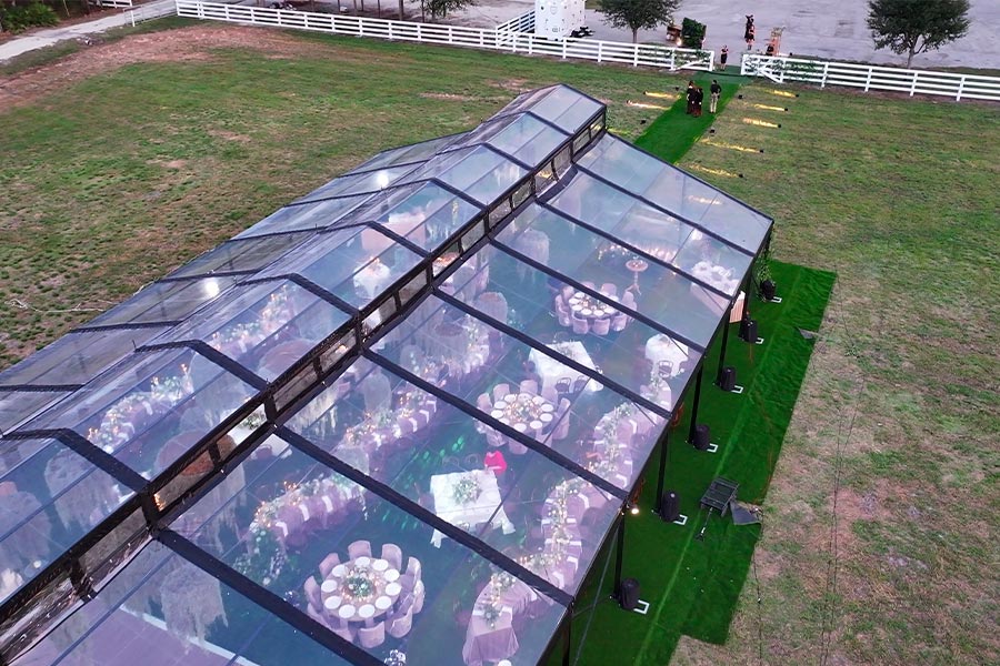 Aerial view of a luxury clear-top wedding tent with elegant reception seating on a green lawn.