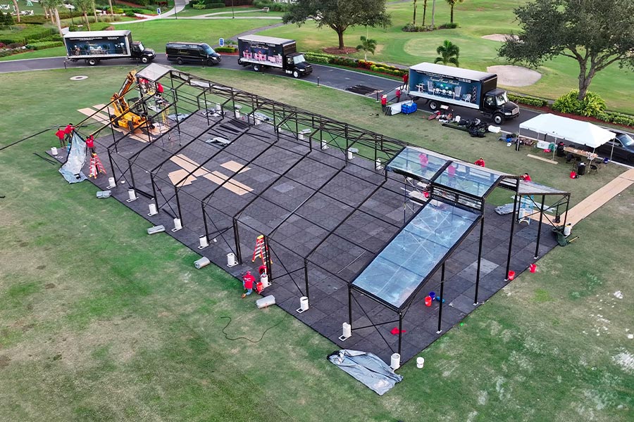 Large scale event production with equipment trucks and a glass-top tent structure under construction.