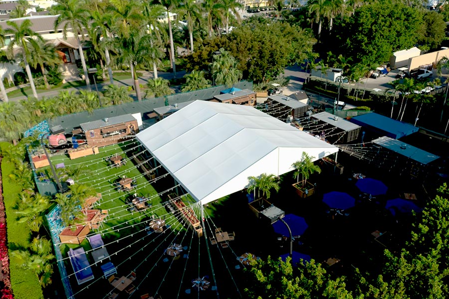 High-angle view of a white event marquee surrounded by string lights, lounge seating, and palm trees.