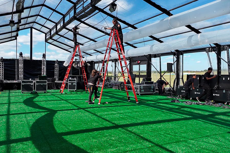 Technicians on ladders installing professional stage lighting and rigging inside a large clear-roof event tent.