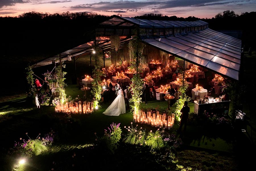 Romantic night wedding reception inside a lit glass tent with candles and hanging greenery.