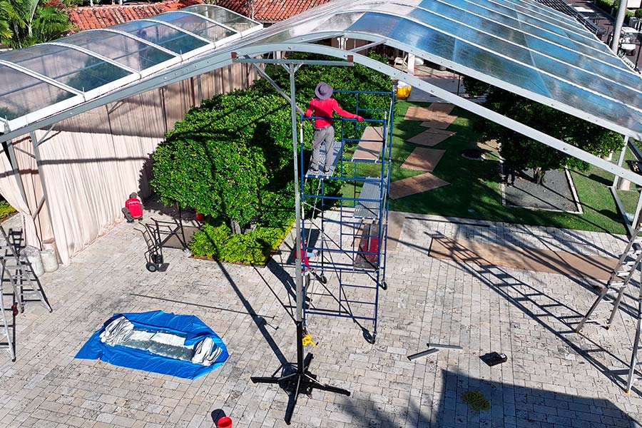 Workers using scaffolding to install a large clear-top frame tent over a paved patio and garden area.