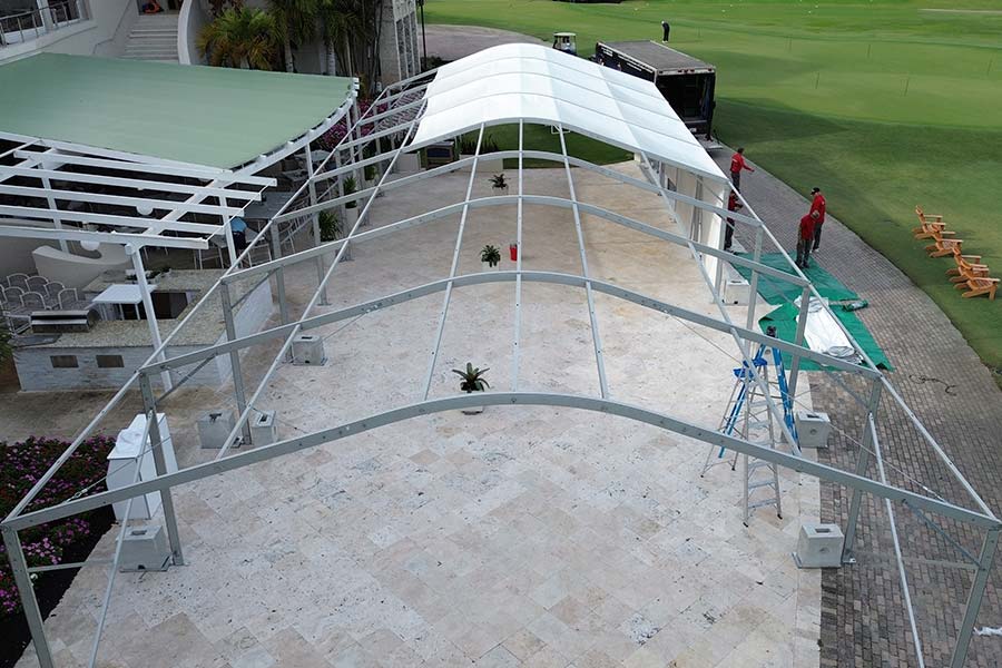 Large clear span tent frame being assembled on a light-colored tiled patio near a white resort building.