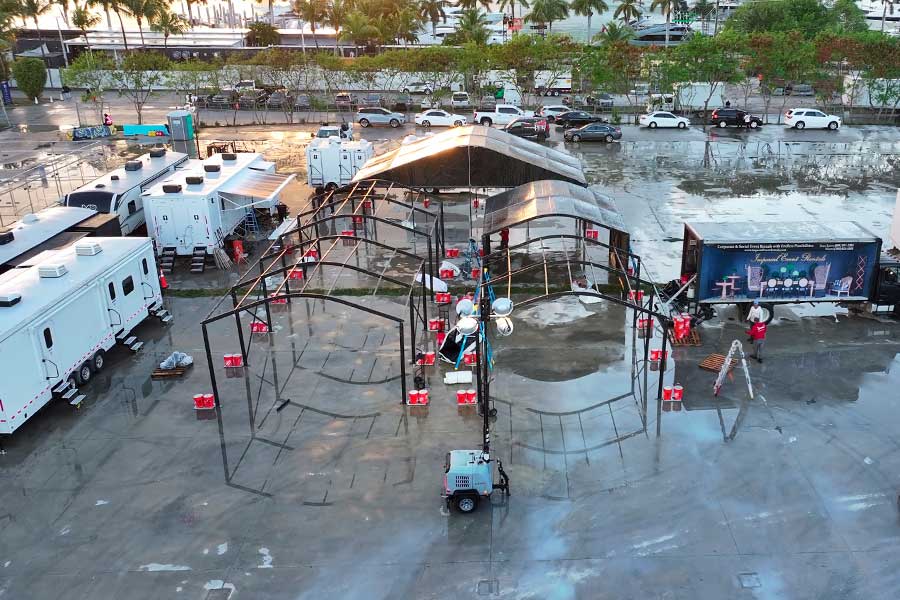 Large frame tent setup in progress on wet paved lot with trailers and vehicles in background.