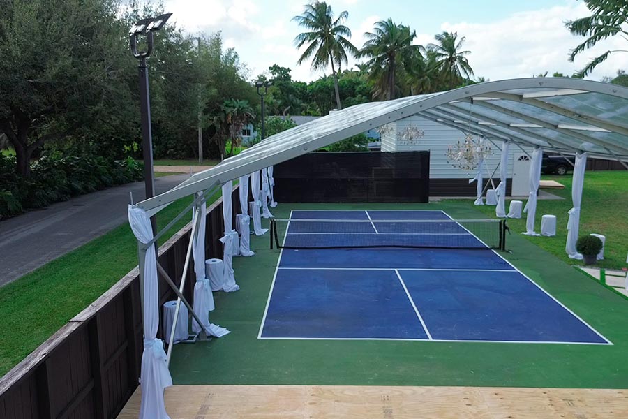 Large clear span tent covering a blue and green outdoor pickleball court with white drapery and chandeliers.
