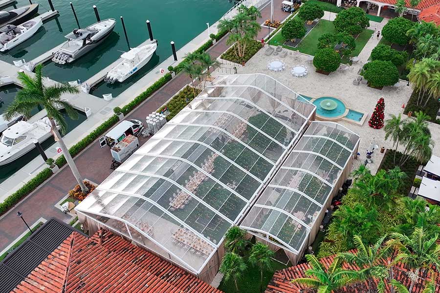 Aerial view of two joined clear wedding tents set up next to a marina and Mediterranean-style building.