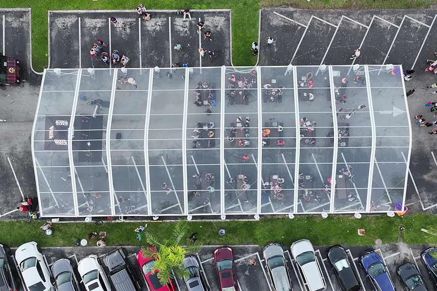 Overhead view of a clear top tent used for a crowded social gathering in a paved parking lot.