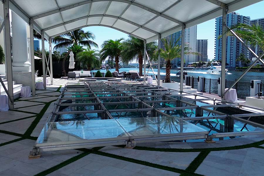 Clear acrylic dance floor over a swimming pool under a white event tent near a waterfront city view.