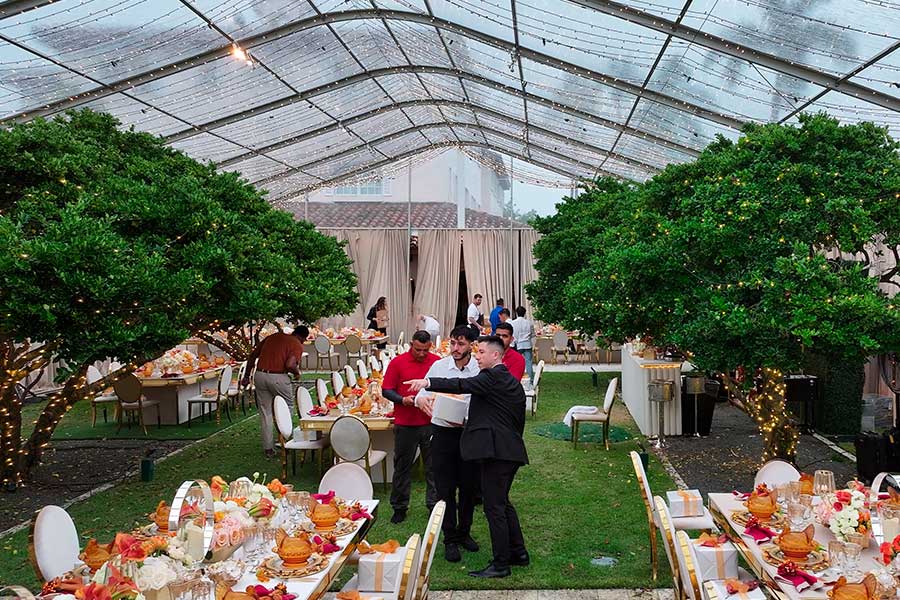 Elegant clear-roof tent interior decorated for a dinner party with trees, long tables, and festive fairy lights.