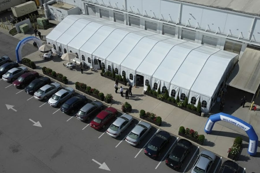 Large white modular frame tent with window sidewalls set up in a loading dock area next to a row of parked cars.