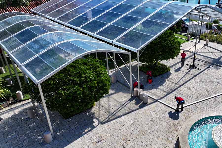 Aerial view of two joined clear-top frame tents being set up over a paved patio and large bushes.