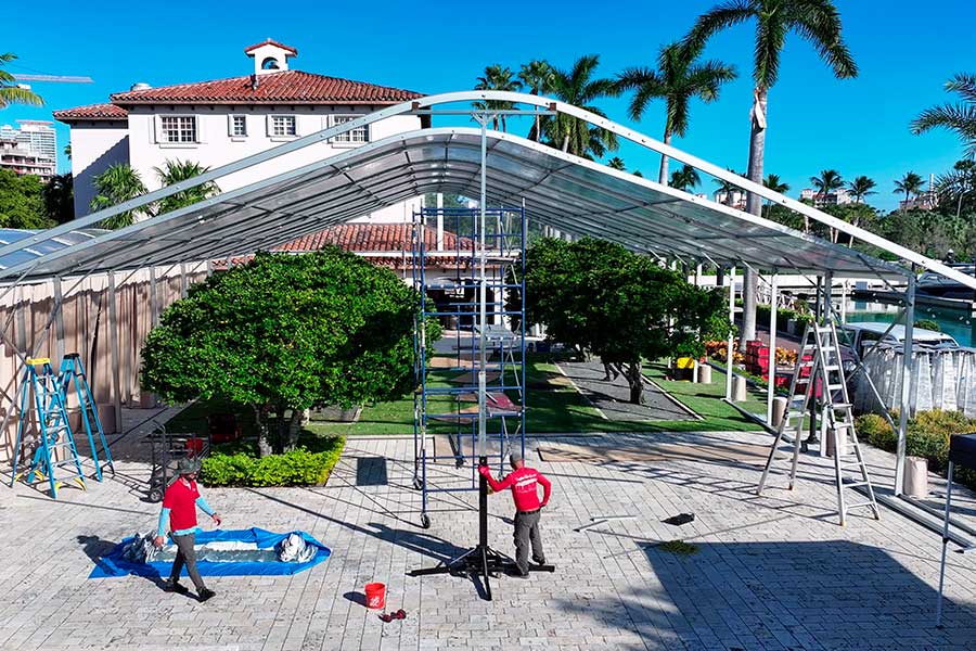 Event crew installing the curved frame of a large clear-span tent on a brick patio next to the water.