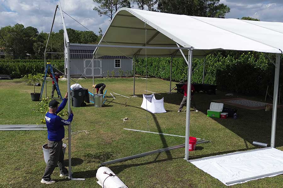 Installation of a white outdoor event tent, showing the metal frame and crew securing the canopy material.