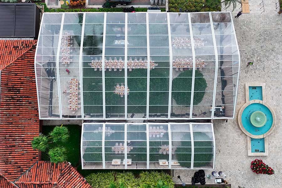 Overhead view of two clear-top event tents covering banquet tables on grass between a building and fountain.