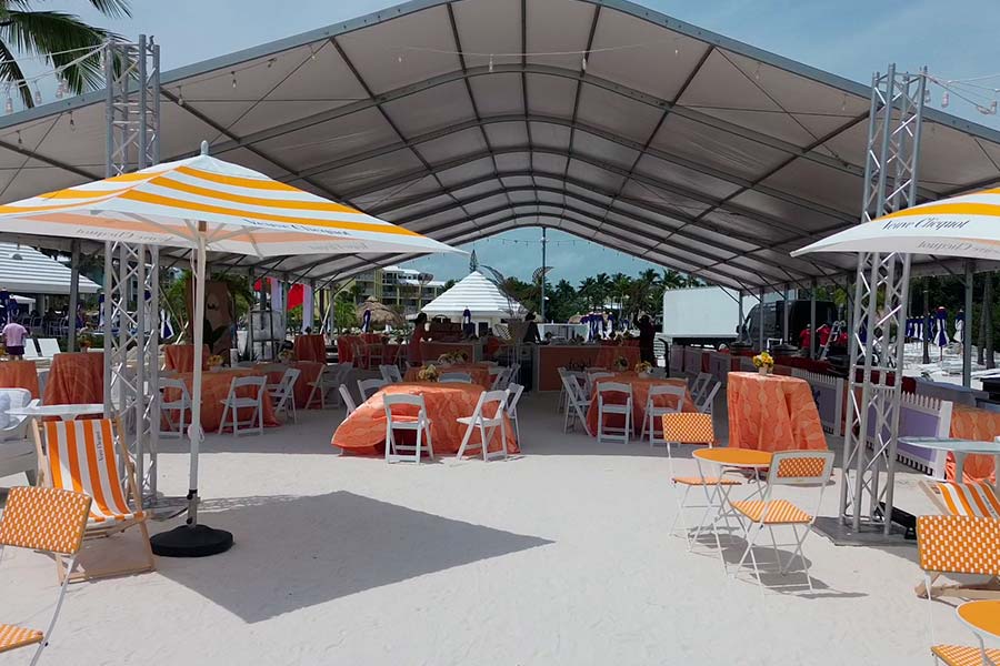 Large white frame tent on a beach with orange tables, white chairs, and branded umbrellas for an event.