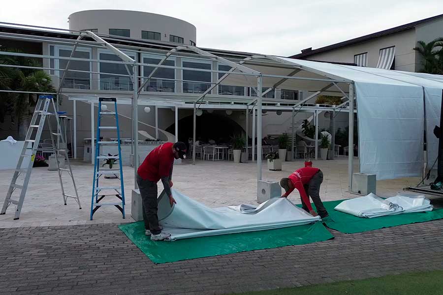 Two workers installing the white canopy on a tent frame setup near a modern outdoor patio.