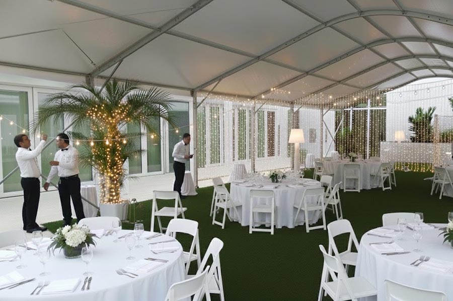 White event tent interior with string lights, round tables, white chairs, and palm décor for a reception.