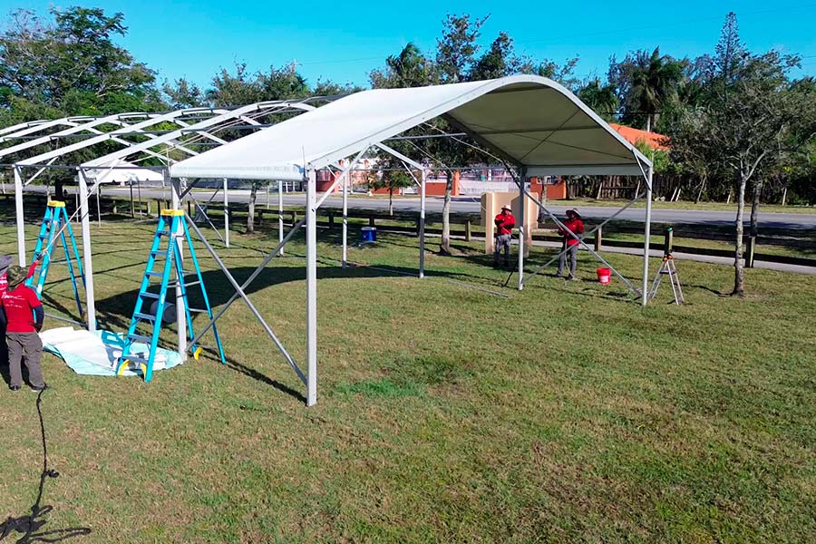 Workers erecting the frame and installing the white top of a large event tent in a grassy field.