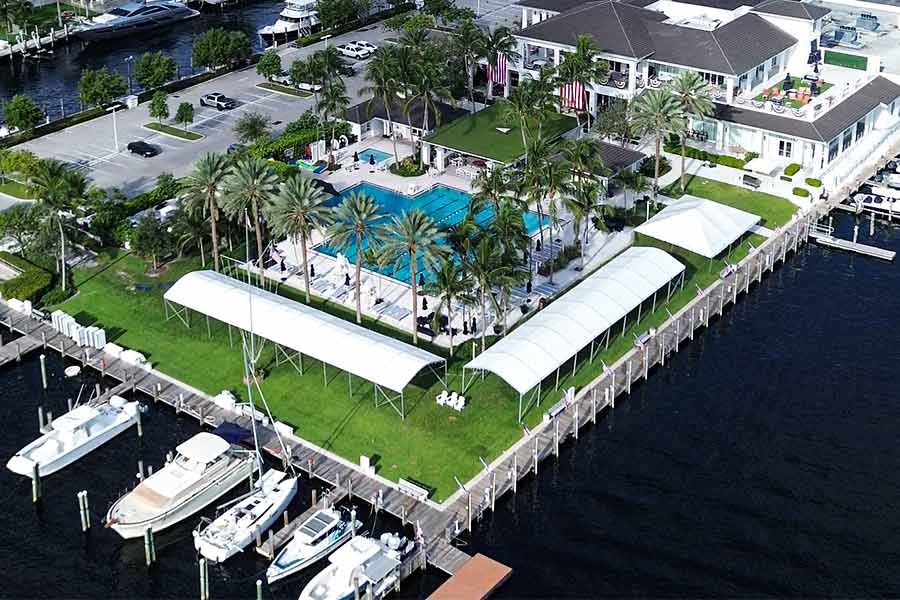 Drone view of waterfront event setup with white canopy tents and yachts near resort pool.