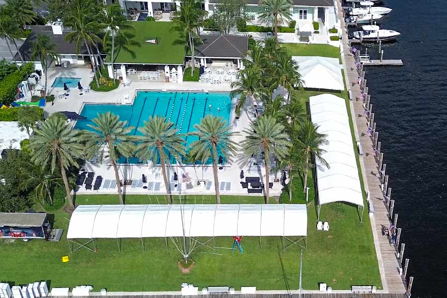 Aerial view of waterfront event setup with white canopy tents beside resort pool and palm trees.