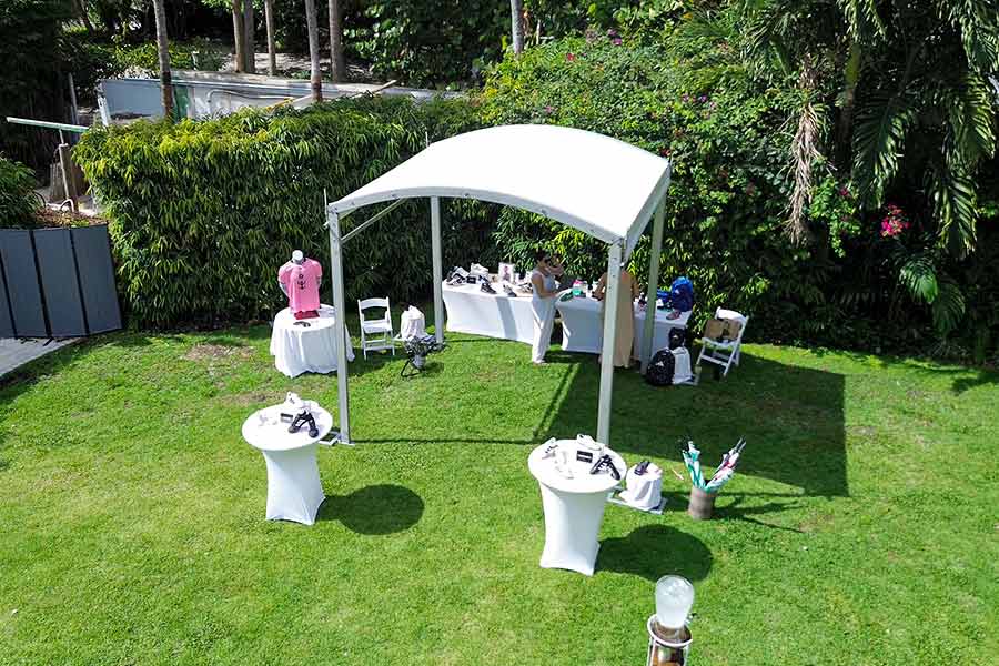 Small white curved tent setup with cocktail tables on lawn surrounded by tropical greenery.