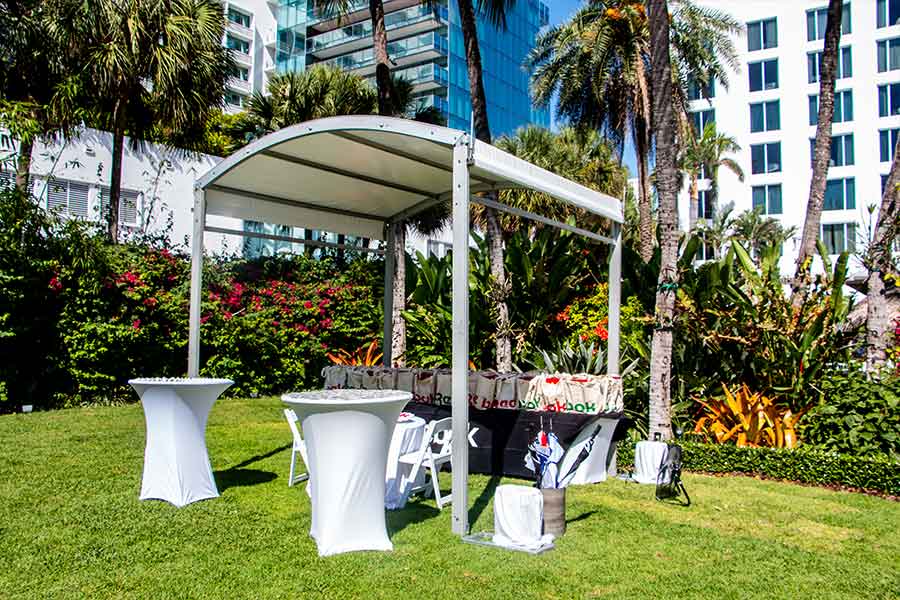 Small white curved tent setup with tall tables for outdoor event under palm trees.