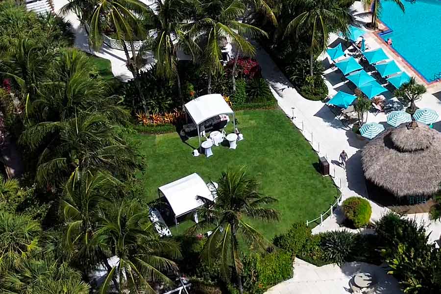 Aerial view of garden event with white canopy tents near poolside and tropical palm trees.