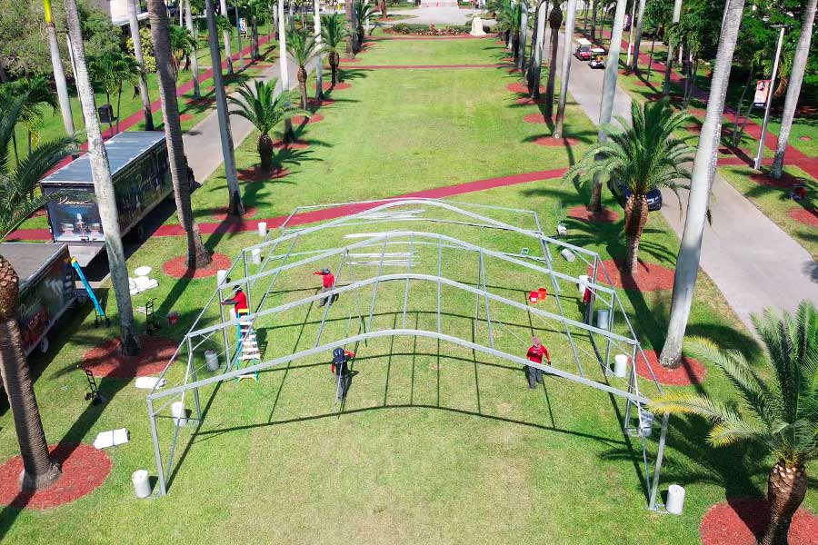 Event team constructing a large tent frame on a long, grassy resort lawn bordered by palm trees.