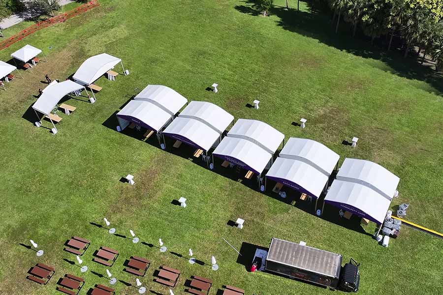 Row of white and purple tents in a park with picnic tables, maximizing comfort and style for a festival.