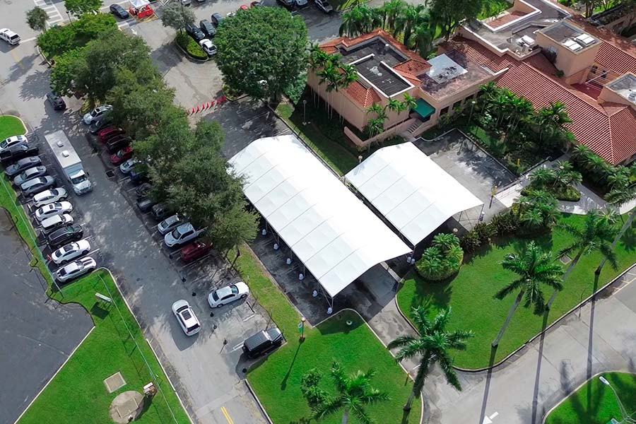 Aerial view of two white frame tents in a parking lot next to a commercial building and palm trees.