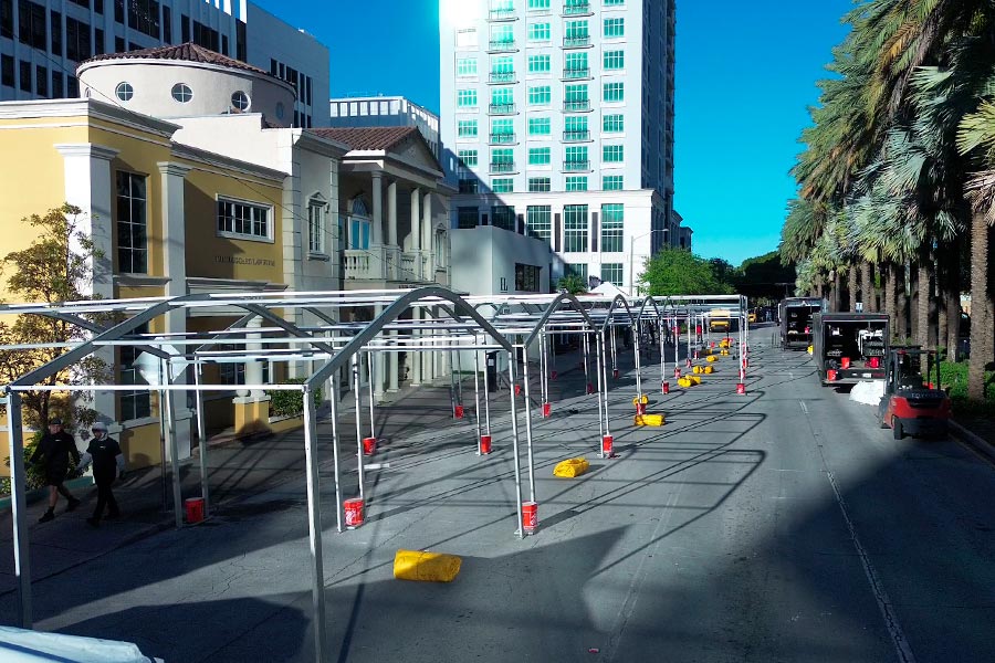 Installation crew setting up the frame of a street event tent in an urban area with tall buildings and palm trees.