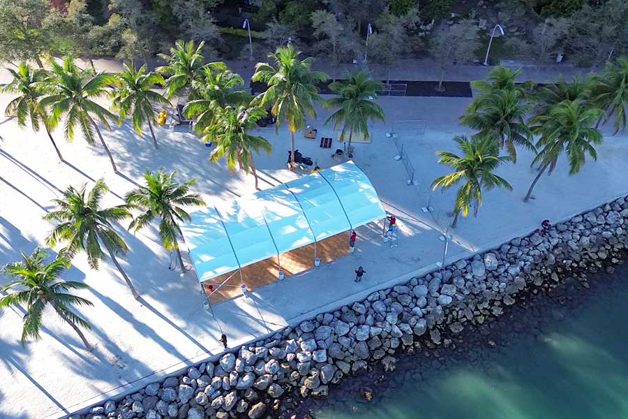 White arched tent with wood flooring on a sandy beach near palm trees and ocean rocks.