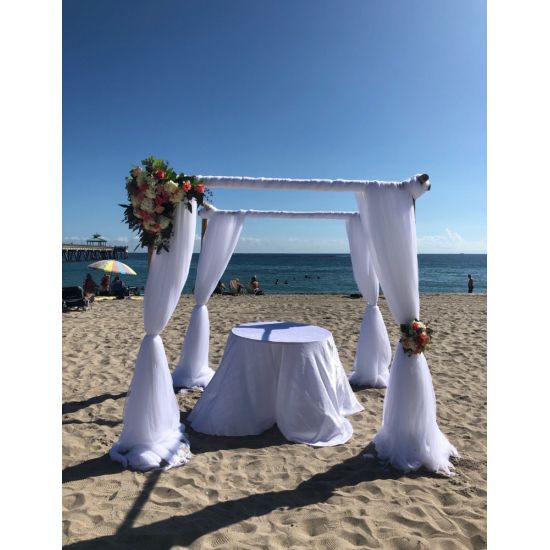 White chuppah canopy on four posts with fabric draping and floral accent, wedding ceremony structure for beach and outdoor events.