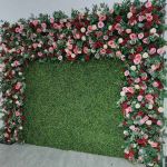 Green hedge wall with floral border frame of pink and white blooms, garden-style backdrop panel for weddings and photo booth setups.